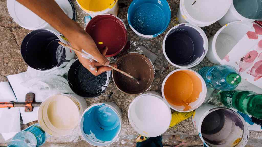 Multiple jars of different colored paint with a person reaching for one with a paintbrush in their hand