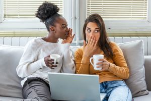 Two women drinking coffee on a couch appearing to gossip