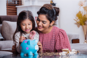 A woman and a child sitting together on the floor placing money into a blue piggy bank