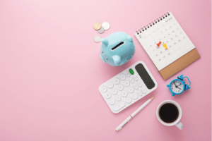 Pink desk with a calendar, calculator, piggy bank, pen, coffee, and clock sitting on top