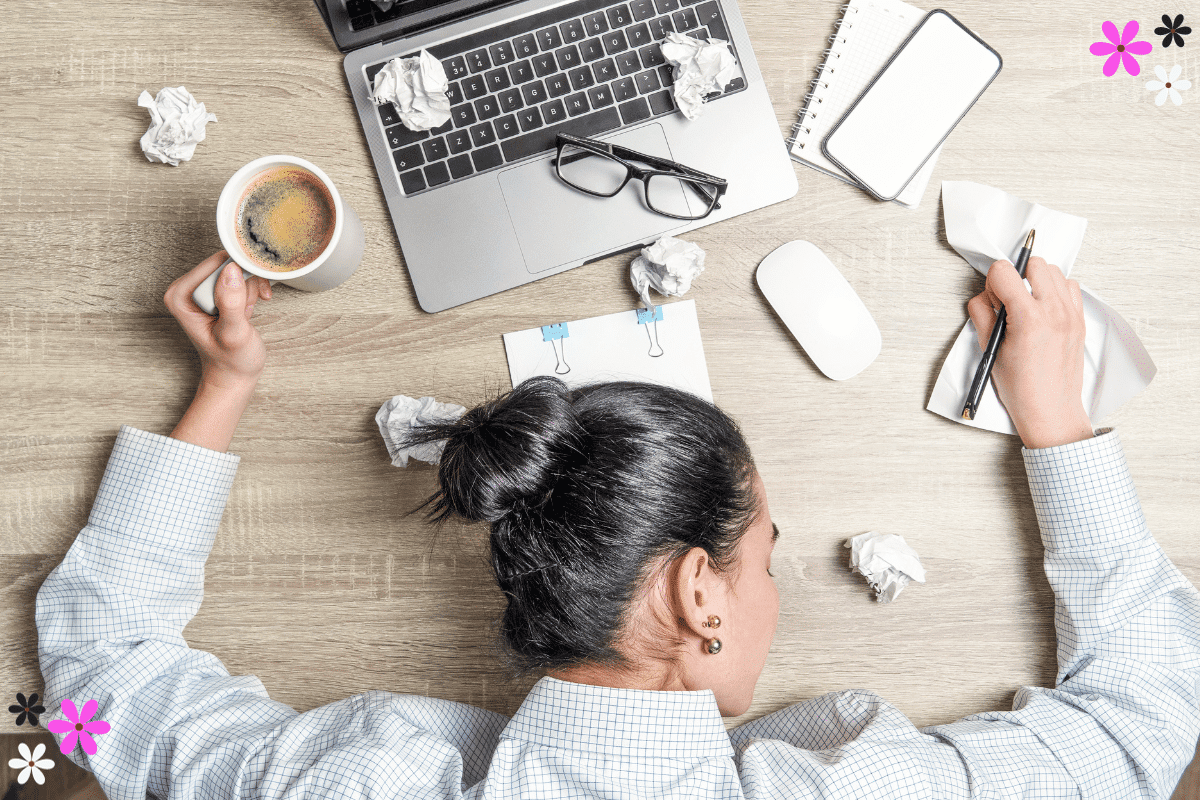 Woman with her head on a desk with many crumbled up papers, a laptop, and coffee